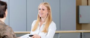 Eine Frau in einem weißen Hemd lächelt und übergibt ein Dokument und eine Fernbedienung an eine andere Person. Der Hintergrund zeigt eine Wand mit einem Bilderrahmen und grauen Schränken.