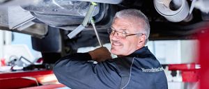 Ein Mann in einer Mercedes-Benz Uniform arbeitet unter einem Auto und benutzt ein Werkzeug, um den Unterboden des Fahrzeugs zu inspizieren oder zu reparieren.
