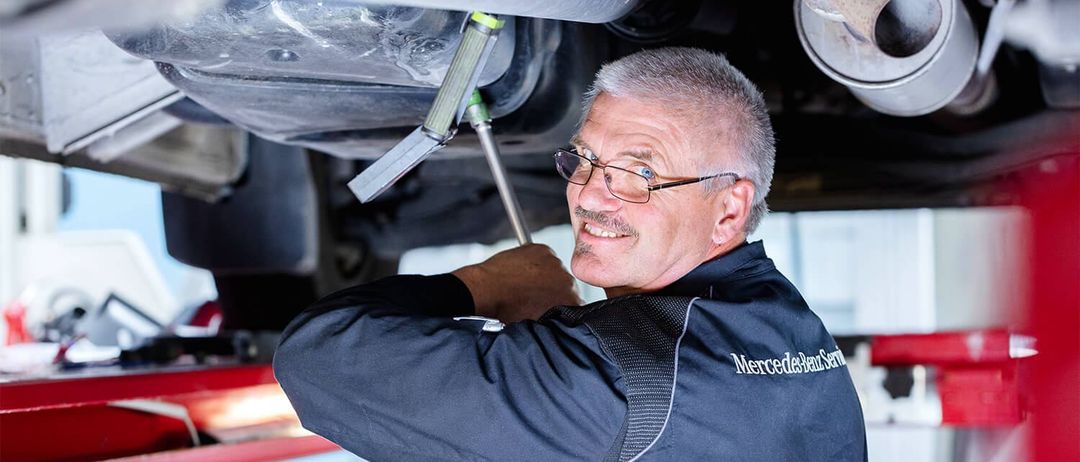 Ein Mann in einer Mercedes-Benz Uniform arbeitet unter einem Auto und benutzt ein Werkzeug, um den Unterboden des Fahrzeugs zu inspizieren oder zu reparieren.