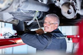 Ein Mann in einer Mercedes-Benz Service-Uniform arbeitet an einem Auto-Unterboden und benutzt ein Werkzeug. Er lächelt und scheint sich auf seine Aufgabe zu konzentrieren.