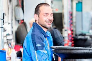 Ein lächelnder Mann in einer blauen Ford-Jacke steht in einer Werkstatt mit einer schwarzen Ölablasspfanne in der Nähe.