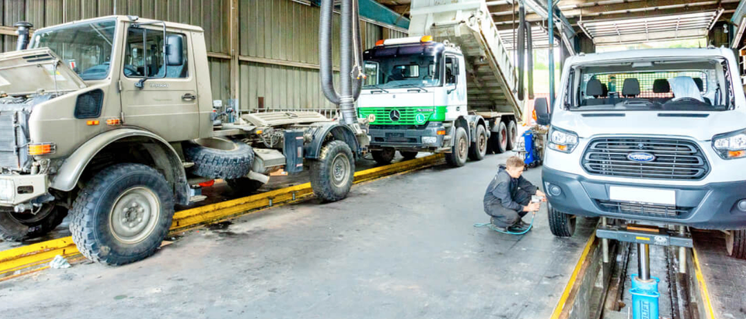 Ein junger Mann arbeitet in einer Werkstatt an einem Fahrzeug, wobei zwei Lastwagen in der Nähe geparkt sind. Ein Lastwagen hat einen großen Auspuff.