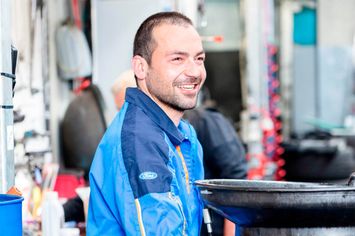 Ein Mann in einer blauen Ford-Jacke lächelt und steht in einer Werkstatt.