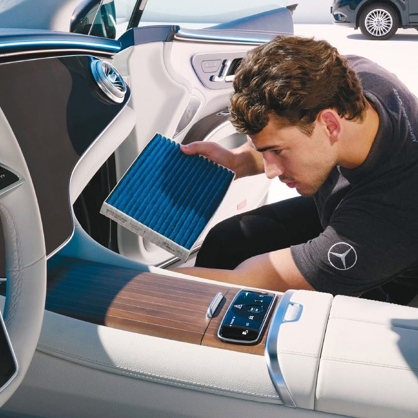 Ein Mann in einem schwarzen Mercedes-Benz-Shirt sitzt auf dem Fahrersitz eines Autos und hält einen blauen Luftfilter in der Hand.