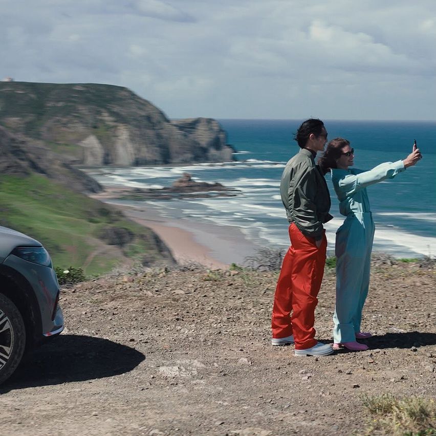 Ein Paar steht auf einem Felsvorsprung mit Blick auf den Ozean, einer macht ein Foto mit dem Handy. Ein Auto ist in der Nähe geparkt.