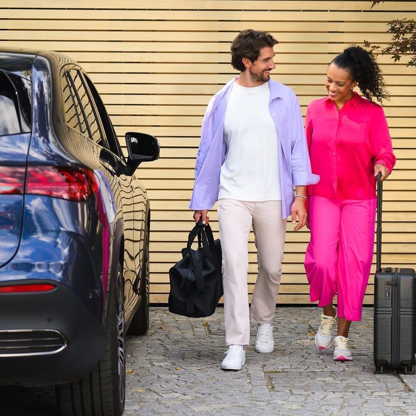 Ein Paar, ein Mann und eine Frau, geht mit Gepäck neben einem blauen Auto auf einem gepflasterten Bereich.