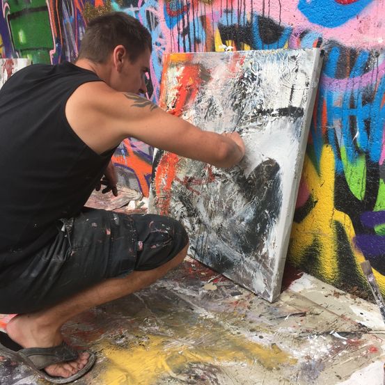 Ein Mann in einem schwarzen Tanktop und Shorts malt eine große Leinwand an einer mit Graffiti bedeckten Wand.
