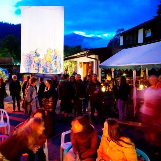 Eine lebhafte Zusammenkunft im Freien bei Nacht mit Menschen, die sich unterhalten, einem großen abstrakten Gemälde auf einem weißen Hintergrund und einem Hintergrund aus Häusern und Bäumen unter einem blauen Himmel.
