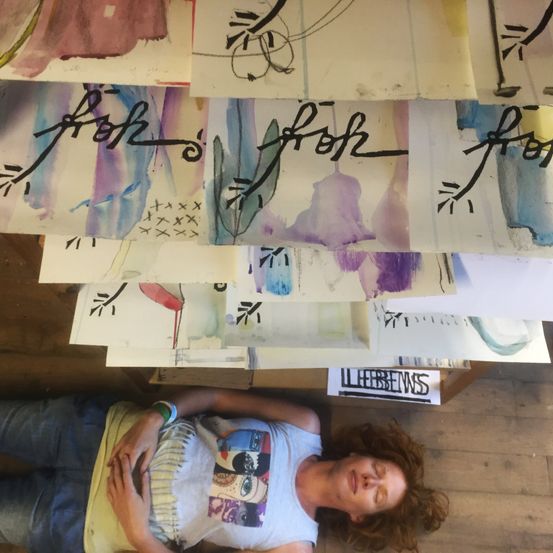Eine Person liegt mit geschlossenen Augen auf dem Boden, umgeben von mehreren Aquarellgemälden, die auf einem Holztisch gestapelt sind. Die Gemälde zeigen abstrakte Designs und Text.