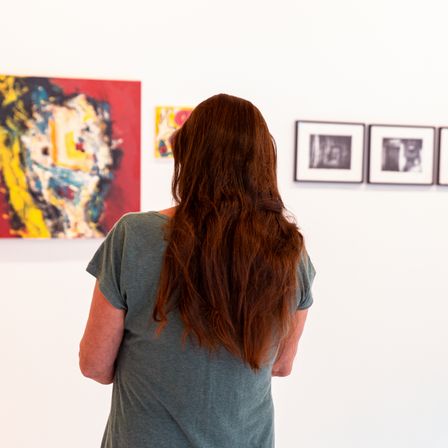 Eine Frau mit langen braunen Haaren steht vor einer weißen Wand mit mehreren gerahmten Kunstwerken. Das größte Gemälde ist abstrakt und farbenfroh, während die anderen schwarz-weiß sind.