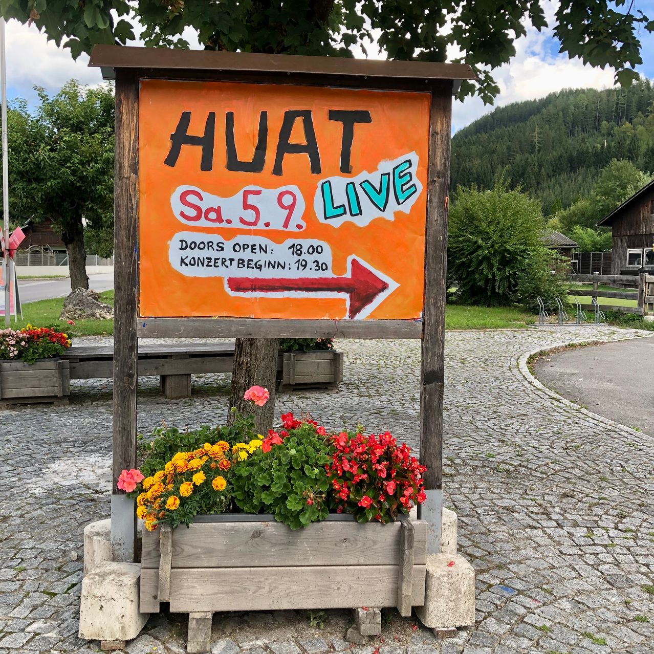 Ein Schild mit orangefarbenem Hintergrund und schwarzem Text steht für 'HUAT Sa.5.9. LIVE'. Das Schild hat einen roten Pfeil, der nach rechts zeigt. Unten am Schild stehen Blumen in einem hölzernen Pflanzkasten. Im Hintergrund befindet sich ein Berg mit Bäumen.