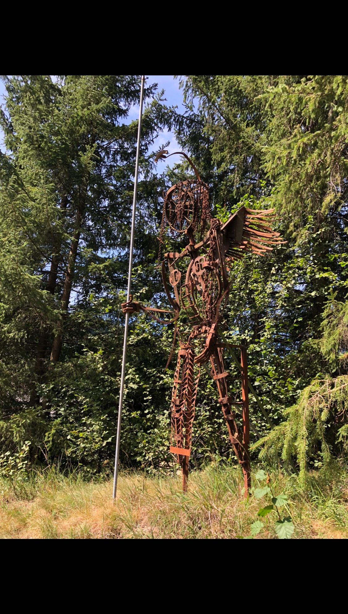 Eine verrostete Metallskulptur einer Figur mit Flügeln und einer Lanze steht in einem Wald.