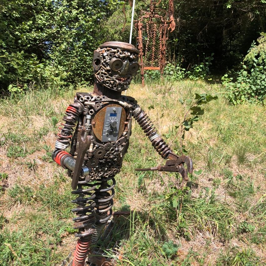 Eine metallische Roboterskulptur steht in einem Grasfeld, hält eine Zange und ist von Bäumen umgeben.