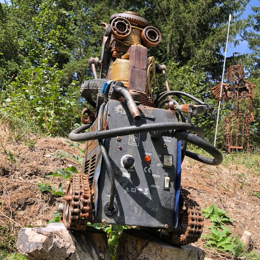 Ein mechanischer Roboter aus alten Maschinenteilen steht auf einem Baumstumpf im Wald.