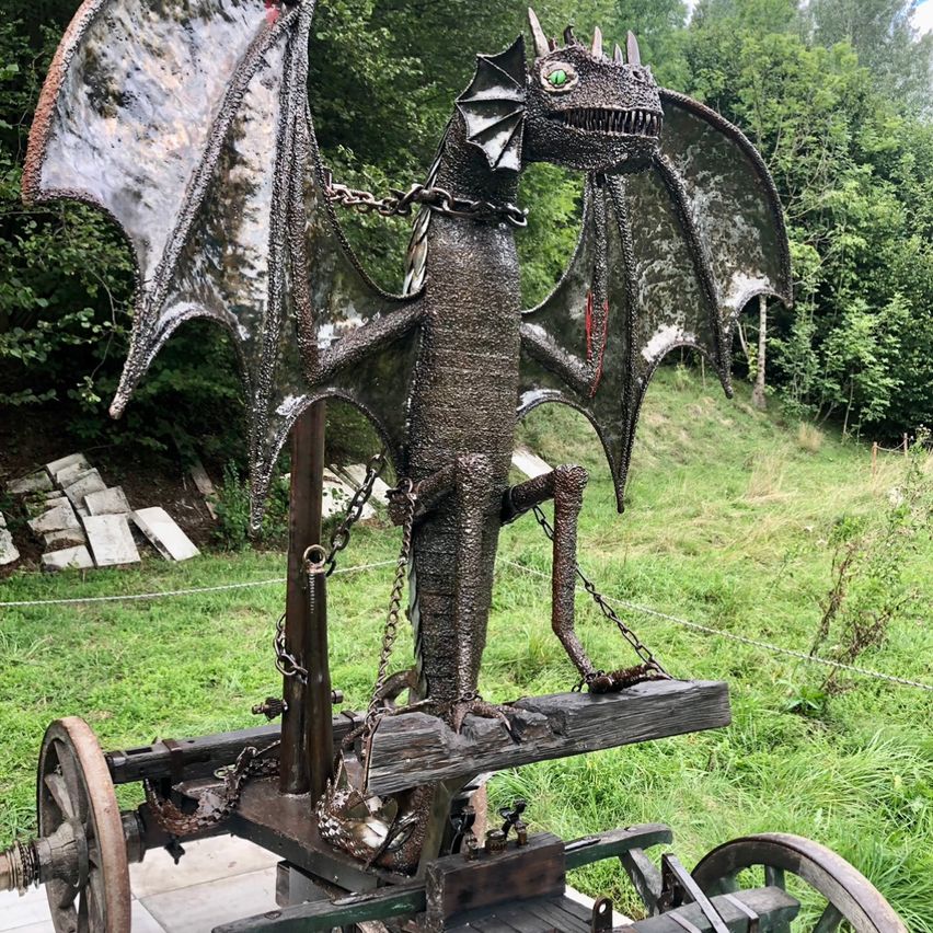 Eine metallische Drachenstatue mit grünen Augen, auf einem hölzernen Wagen mit Ketten, vor einem grünen Hintergrund mit Bäumen.
