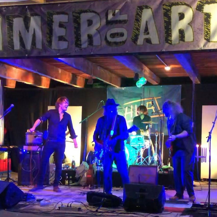 A band performs on a stage with a banner that reads 'Summer's Art'. The lead singer plays guitar while the drummer and another guitarist play behind him. The stage is lit with blue lights.