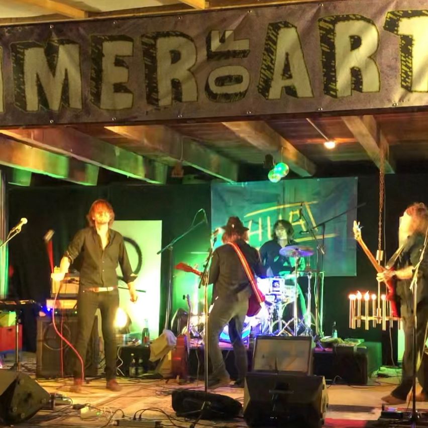 A band performs on stage with a banner reading 'Summer of Art' above them. The stage is lit with green and yellow lights.