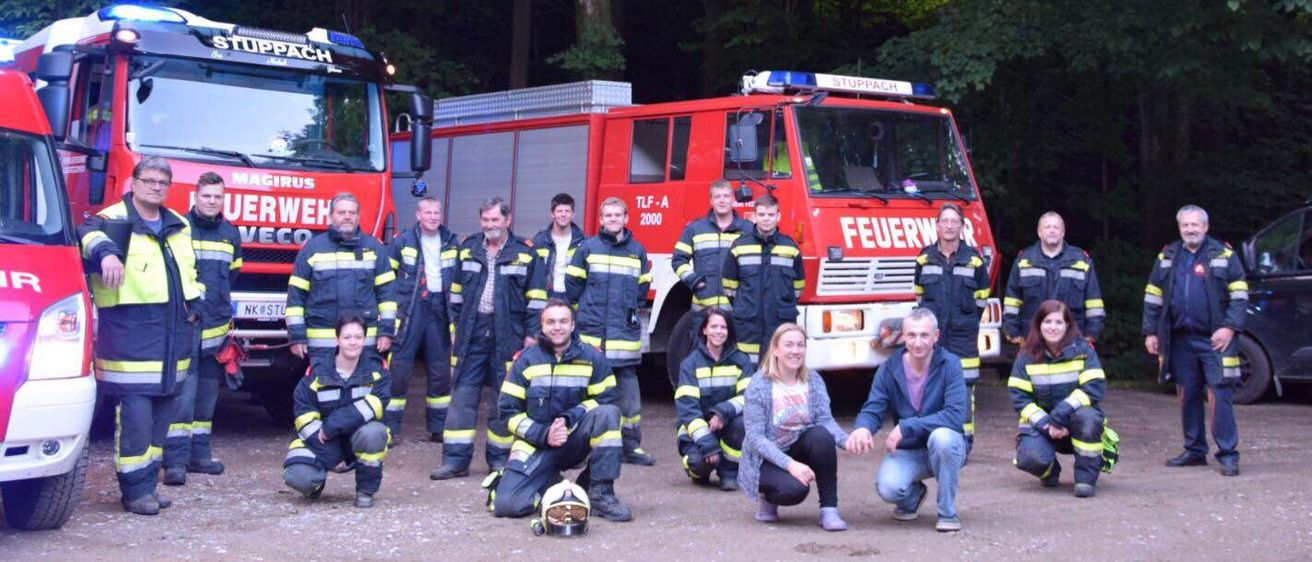 Eine Gruppe von Feuerwehrleuten und ihren Familien posiert für ein Foto vor Feuerwehrautos in einem bewaldeten Gebiet.