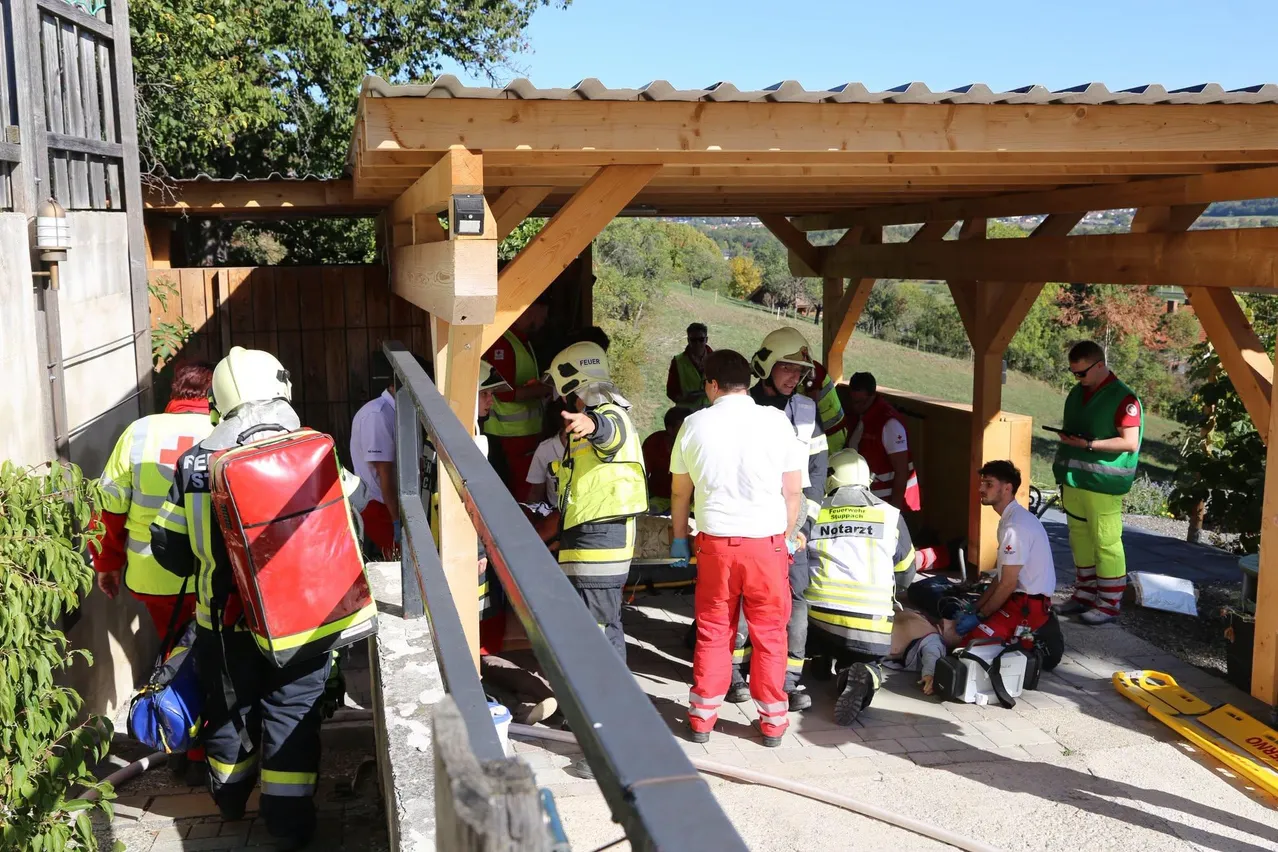 Eine Gruppe von Feuerwehrleuten und medizinischem Personal versammelt sich um eine Person, die auf einer Trage unter einem hölzernen Pavillon liegt.