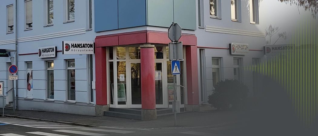 Ein Ladengeschäft mit einem Schild, das 'HANSATON Horsysteme' auf einem mehrstöckigen Gebäude anzeigt. Es gibt Glastüren und Fenster sowie ein blaues Fußgängerübergangsschild.