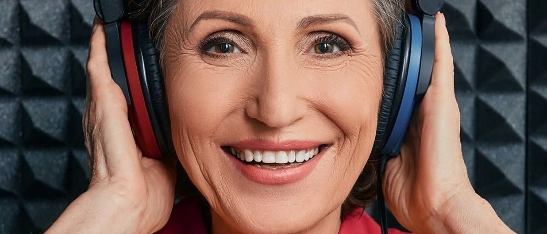 Eine ältere Frau mit einem roten Hemd lächelt und hält Kopfhörer am Kopf. Der Hintergrund ist schwarz-weiß. Es gibt einen grünen Kreis mit weißem Text.