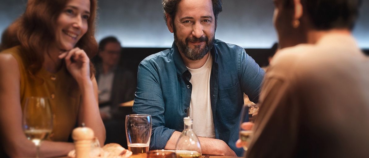 Ein Mann und eine Frau lächeln sich in einem Restaurant an. Der Tisch ist mit Tellern, Weingläsern, einer Ölflasche und einem Korb mit Brot gedeckt. Eine weitere Person sitzt im Hintergrund.