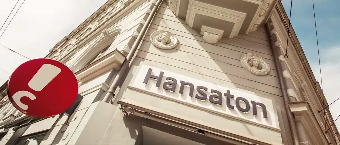 Ein weißes Gebäude mit Hansaton auf einem Schild. Dekorationen mit Büsten sind über dem Schild. Ein rotes Objekt ist links.