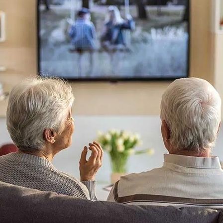 Zwei ältere Personen sitzen auf einem Sofa und schauen fern. Der Mann trägt ein weißes Hemd, die Frau einen grauen Pullover. Ein Fernseher zeigt eine Familie.