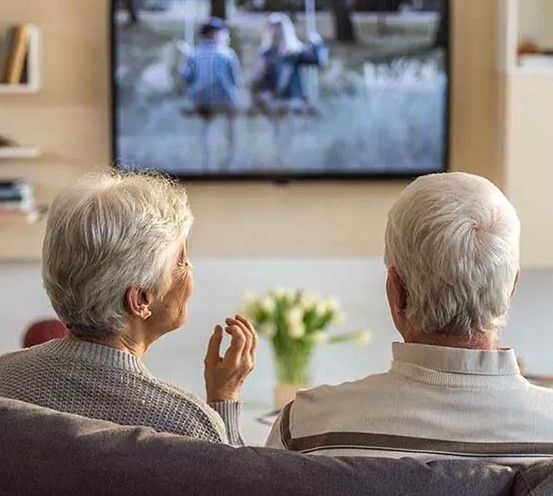 Zwei ältere Personen sitzen auf einem Sofa und schauen fern. Der Mann trägt ein weißes Hemd, die Frau einen grauen Pullover. Ein Fernseher zeigt eine Familie.