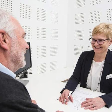 Ein älterer Mann und eine Frau mit Brille sitzen vor einem Schreibtisch, möglicherweise in einem Büro. Die Frau lächelt und hält einen Stift und Papier.