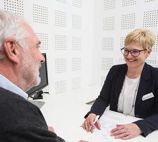 Ein älterer Mann und eine Frau mit Brille sitzen vor einem Schreibtisch, möglicherweise in einem Büro. Die Frau lächelt und hält einen Stift und Papier.