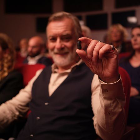 Ein älterer Mann in einer Weste hält ein kleines schwarzes Objekt in einem Theater, um ihn herum sitzen andere Menschen.