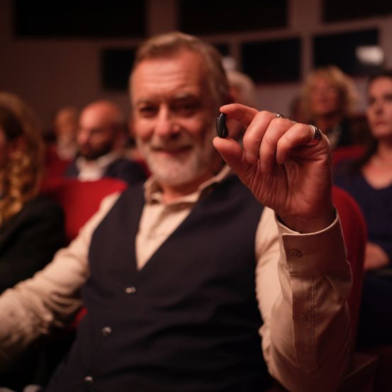 Ein älterer Mann in einer Weste hält ein kleines schwarzes Objekt in einem Theater, um ihn herum sitzen andere Menschen.