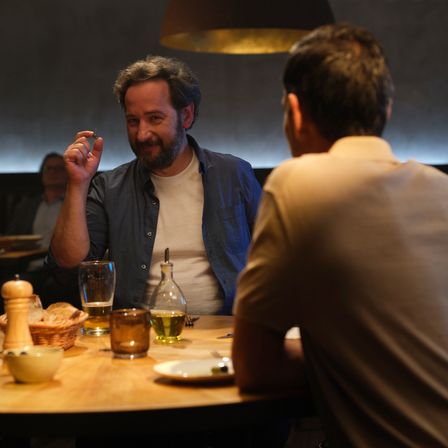 Zwei Männer sitzen an einem Tisch, einer hält ein kleines Objekt, während sie sich unterhalten, eine Lampe darüber, Teller und Getränke.