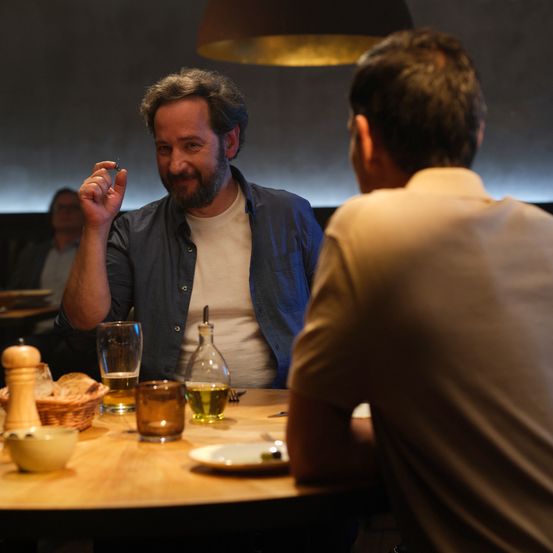 Zwei Männer sitzen an einem Tisch, einer hält ein kleines Objekt, während sie sich unterhalten, eine Lampe darüber, Teller und Getränke.