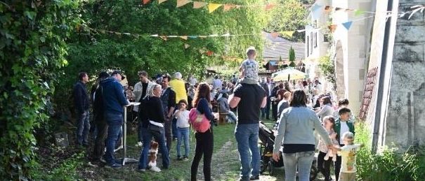 Ein Bild von einem Frühlingsfest mit Menschen in einem Gartenbereich. Der Text auf Deutsch lautet 'Wulkakaistl Fruhlingsfest'. Datum ist 3. Mai, Beginn um 14:00 Uhr. Ort ist Achazium, Forchtenstein. Eine englische Übersetzung ist 'Ein Fest für Genießer'.