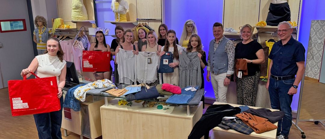Eine Gruppe von Menschen posiert mit Kleidungsstücken in einem Geschäft. Im Hintergrund befinden sich Mannequins und Kleiderständer.