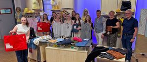Eine Gruppe von Menschen posiert mit Kleidungsstücken in einem Geschäft. Im Hintergrund befinden sich Mannequins und Kleiderständer.
