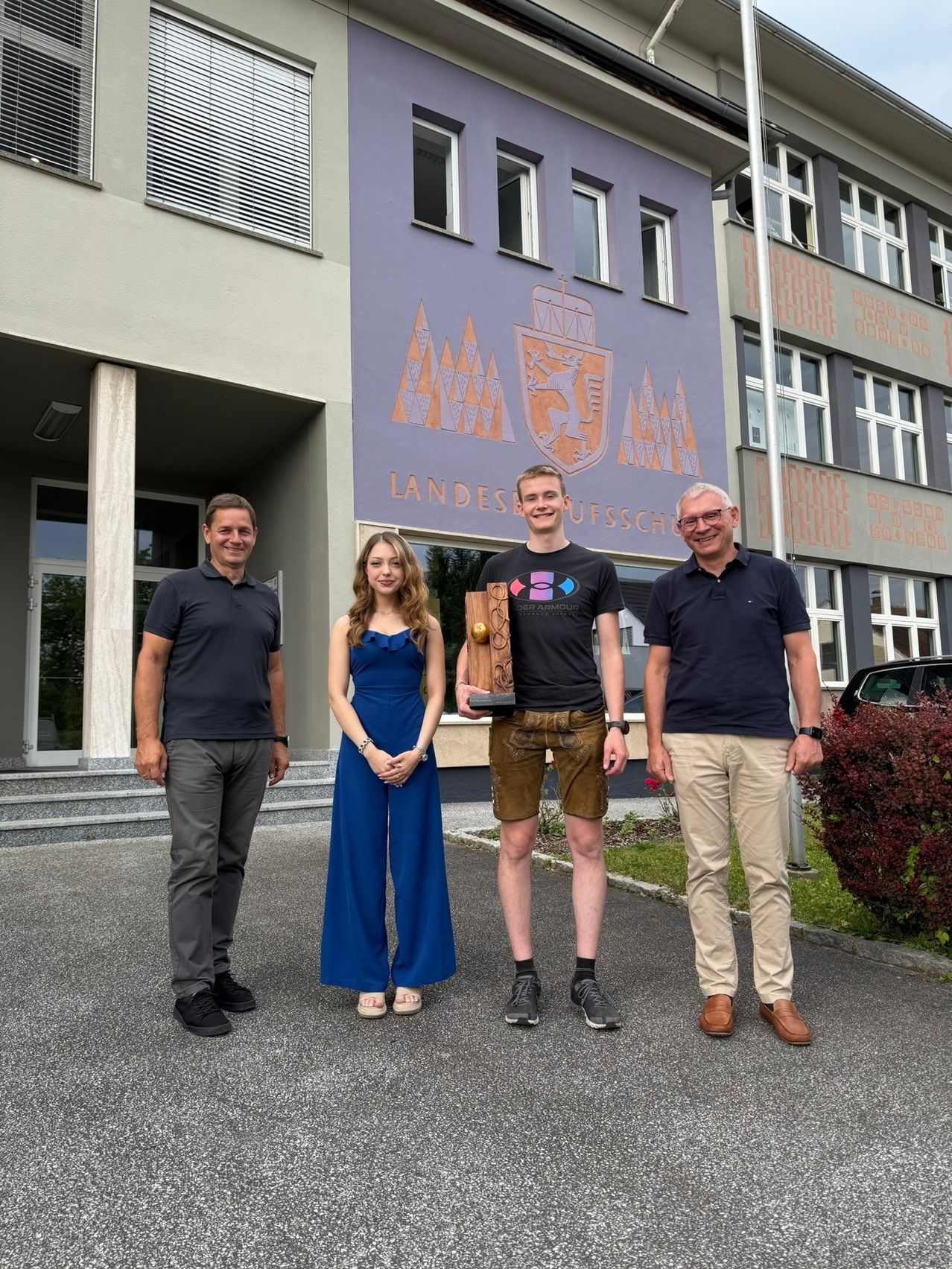 Vier Personen stehen vor einem Gebäude mit einem Wandbild und lächeln für ein Foto. Der Mann auf der rechten Seite hält eine Trophäe.