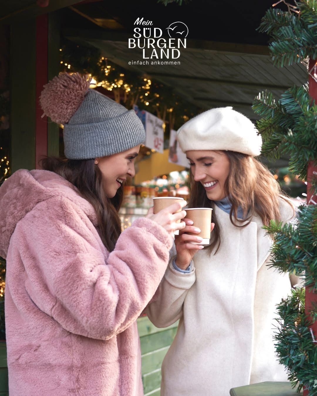 Two women in winter attire, one in a pink coat and the other in a white beanie, holding coffee cups and smiling at a Christmas market.