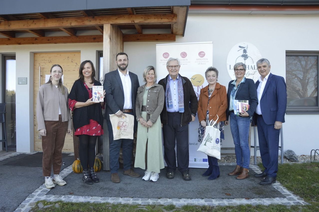 A group of people, including adults and children, are posing for a photo in front of a building. They are holding books and bags. The building has wooden pillars and a signboard with the text Lebensart.