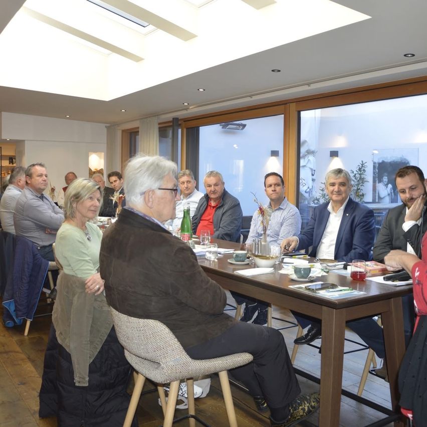 Several people are seated around a table in a dining area, with several items on the table. Some are wearing glasses, while others are wearing sneakers. One of them is wearing a necklace. A woman is standing near the table.