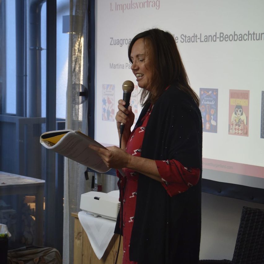 A woman smiles while speaking into a microphone. She holds a paper in one hand and stands in front of a projector screen.