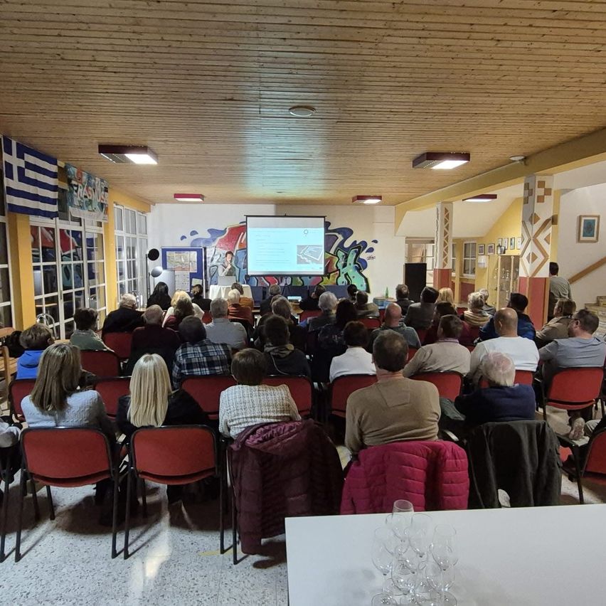 Ein Innenbereich mit Holzdecke. Eine Projektionsleinwand ist an der Wand mit einer Person davor. Eine Menschenmenge sitzt auf roten Stühlen. Auf dem Tisch stehen Weingläser.