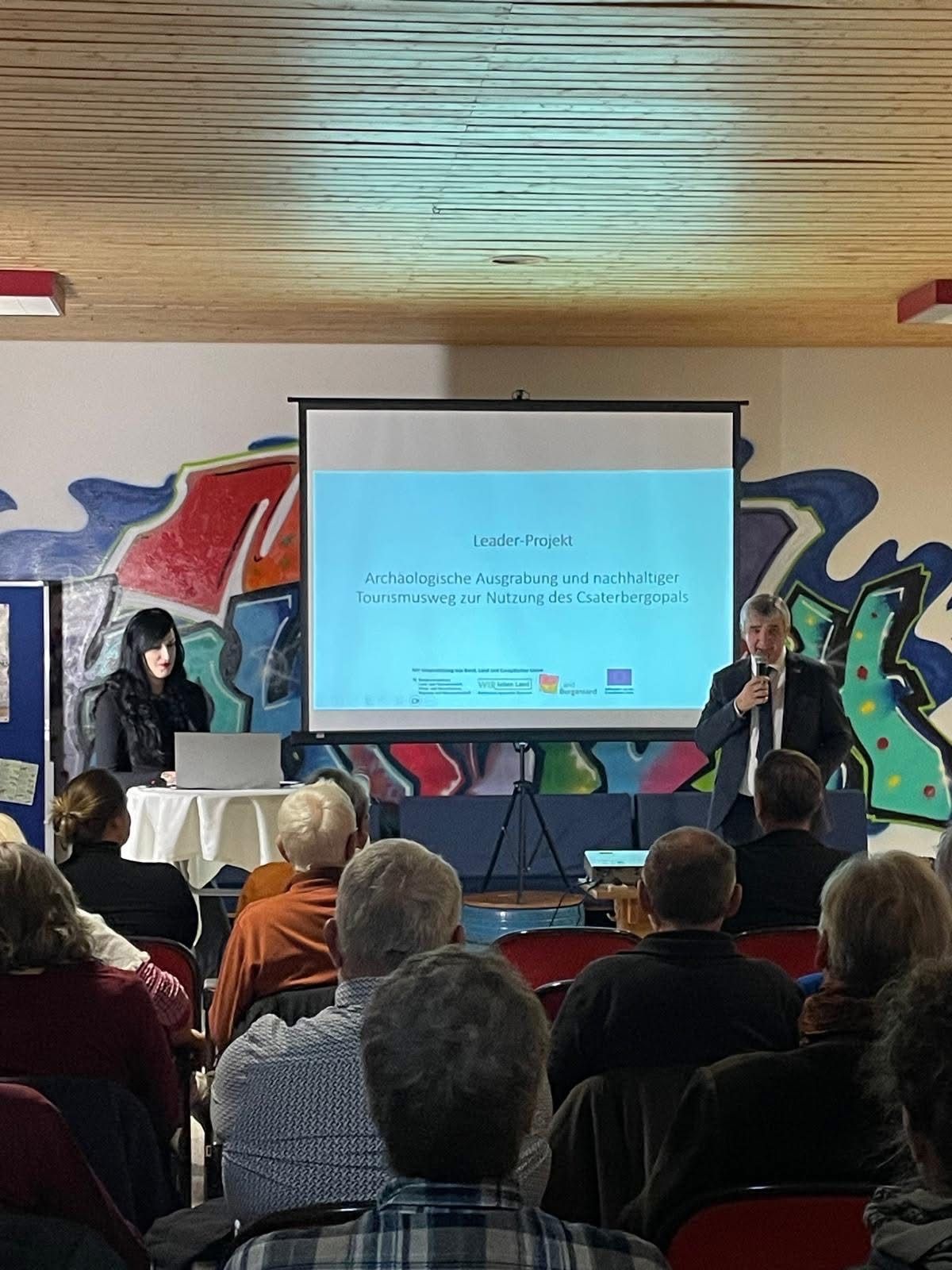 Ein Mann spricht in ein Mikrofon auf einer Bühne vor einem Publikum, während eine Frau an einem Tisch mit einem Laptop sitzt. Eine Projektionsleinwand zeigt eine Präsentation an.