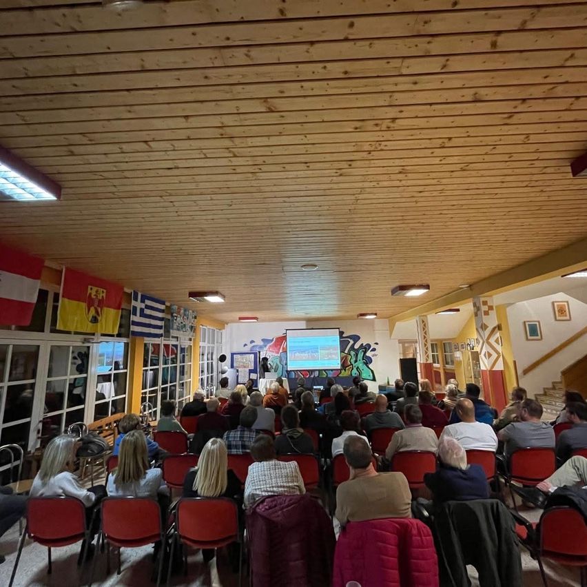 Ein Raum mit Holzdecke, Flaggen an der Wand und einer Projektionsleinwand vorne. Die Menschen sitzen in Reihen und beobachten eine Präsentation. Der Raum ist gut beleuchtet mit Deckenleuchten.