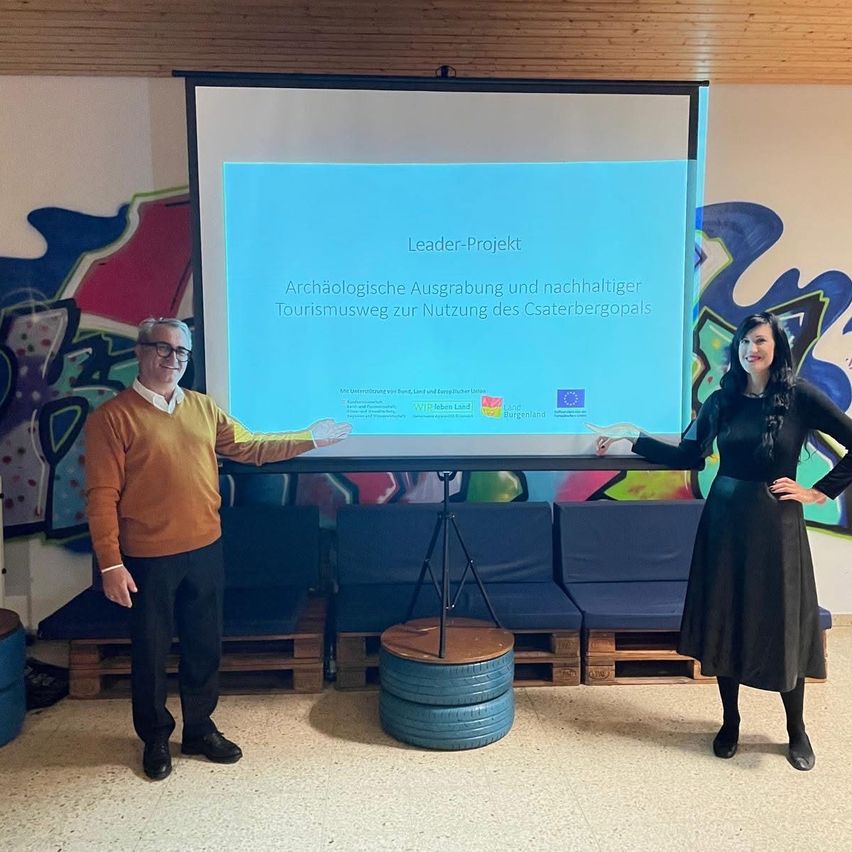 Ein Mann und eine Frau stehen vor einer Projektionsleinwand und zeigen auf ein Bild mit Text über archäologische Ausgrabung und nachhaltigen Tourismus.