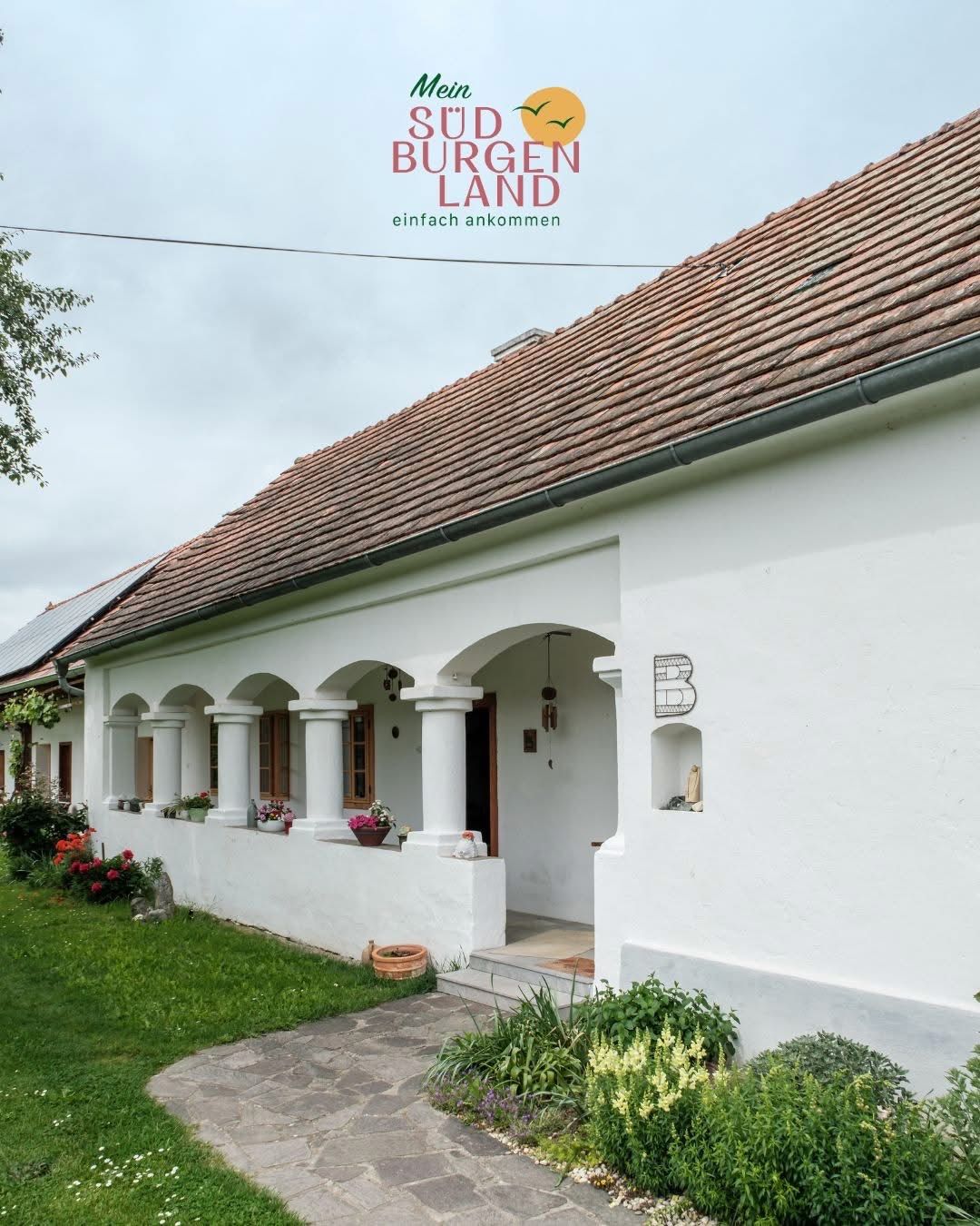 Ein weißes Gebäude mit einem braunen Dach und gewölbten Fenstern steht auf einem grünen Rasen, mit einem Weg, der zum Eingang führt.