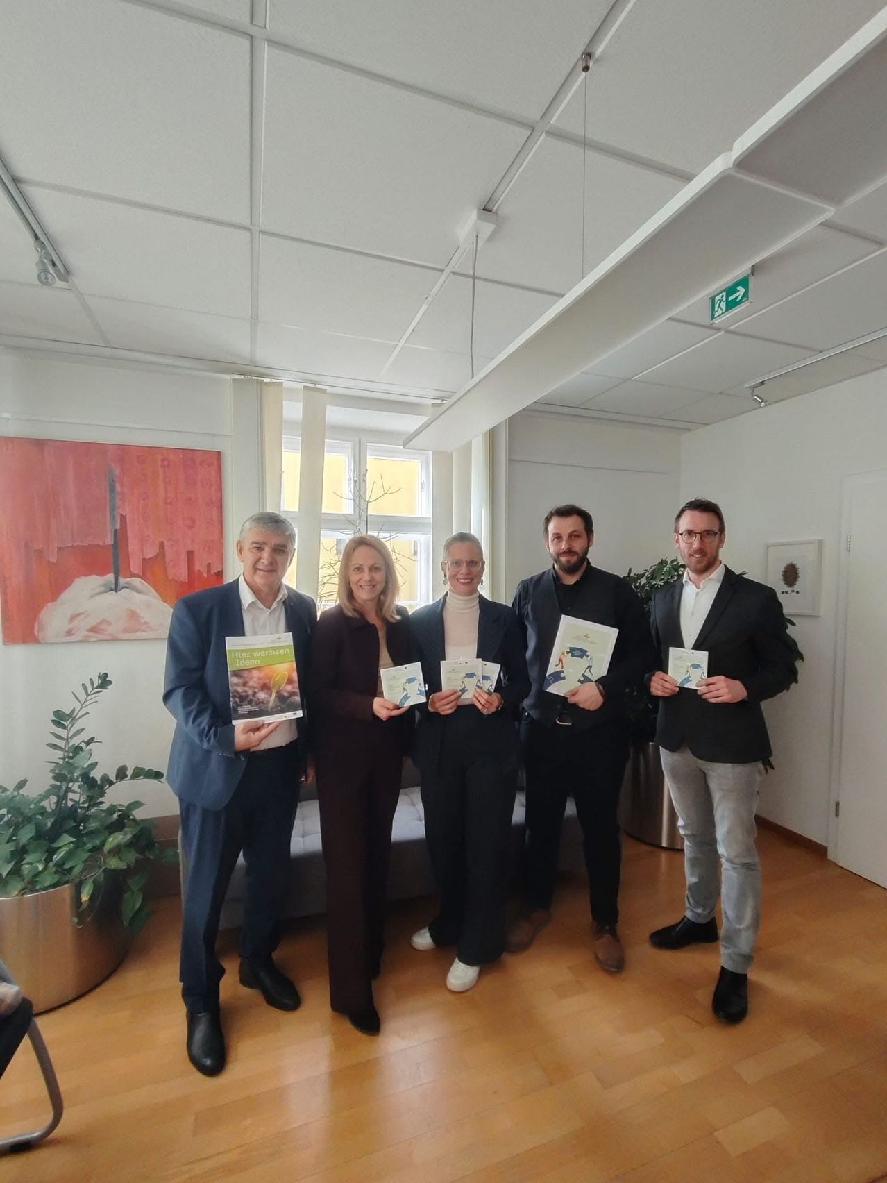 Five people stand indoors, holding booklets, smiling. Behind them, a painting and a window. A potted plant on the left, an exit sign above.