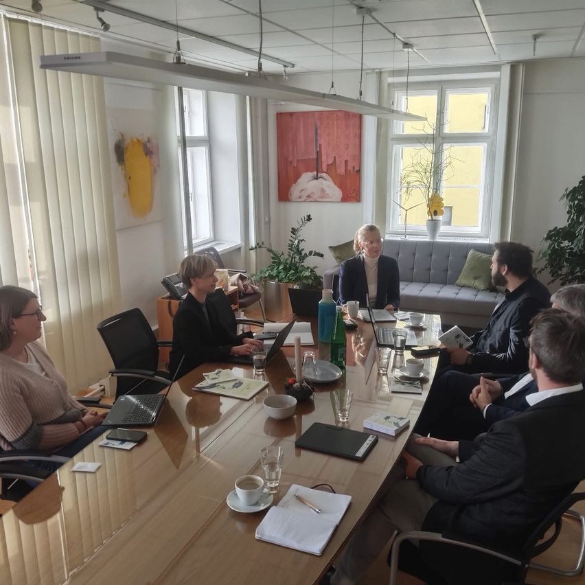 A group of people are gathered around a table in a meeting room, using laptops. Two women and three men are present. The room has white walls and ceilings, with paintings and plants as decorations.